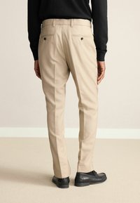 Beige Hosen mit glatter Textur, ausgestattet mit Gesäßtaschen und schwarzen Knopfakzenten. Tragen mit schwarzen Schuhen vor einem neutralen Hintergrund.