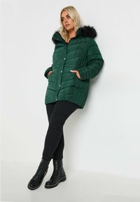 Vadderad grön parkas med huva av päls, med knappstängning, sidofickor och kviltad textur. Svarta byxor och stövlar fulländar looken.