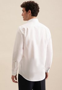 Biała koszula z długim rękawem o gładkiej fakturze, z klasycznym kołnierzykiem i zapięciem na guziki przy mankietach. Back hem jest lekko zaokrąglony.