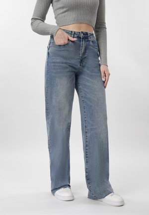 Femeie purtând blugi evazați cu talie înaltă, albastru deschis, pulover scurt gri cu dungi, adidași albi, cu o mână în buzunar și fundal neutru.