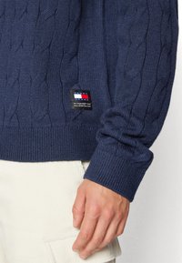 Manche d'un pull en maille torsadée bleu marine avec un patch logo Tommy Jeans au-dessus du bord-côte, pantalon en velours blanc et une main détendue sur le côté.