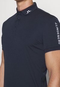 Marinblå polo skjorta med en enfärgad design, knäppt krage och ribbade ärmslut. Har vita logodetaljer på kragen och vänster ärm.