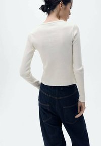 Rippentop in Creme mit langen Ärmeln und tiefem Rückenausschnitt, kombiniert mit dunkelblauen, hochtaillierten Denim-Hosen. Auffällige Nähte an den Taschen.