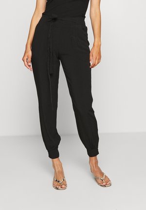 Kobieta ubrana w czarne joggery z wysokim stanem, ściągane przy kostkach i zawiązanym paskiem, w połączeniu z klapkami w wężowy wzór, stojąca na szarej podłodze.