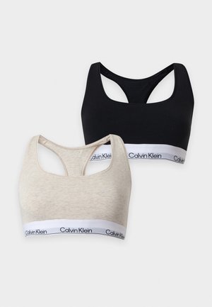 Femme debout pieds nus portant un soutien-gorge de sport noir Calvin Klein et un slip taille haute assorti sur un fond clair uni.