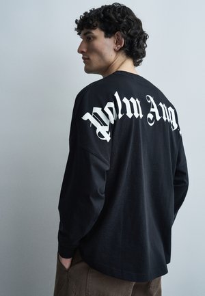 Νεαρός άνδρας με σγουρά μαλλιά που φοράει μαύρο πουκάμισο με μακριά μανίκια και άσπρο γοτθικό κείμενο στην πλάτη, στέκεται πλαγίως μπροστά σε απλό γκρι φόντο.