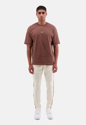 Braunes Baumwoll-T-Shirt mit kurzen Ärmeln und Rundhalsausschnitt, kombiniert mit weißen Hosen mit braunen Seitenstreifen. Getragen mit weißen Sneakern.