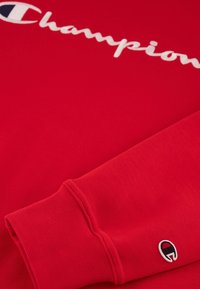 Sudadera roja con el logotipo blanco de "Champion", primer plano del puño con un pequeño logotipo bordado. Textura suave, puños acanalados.