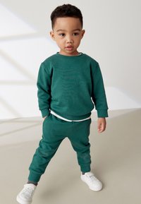 Grüner Baumwoll-Sweatshirt und passende Jogginghosen mit Rippbündchen, kombiniert mit einem weißen T-Shirt und weißen Sneakern.