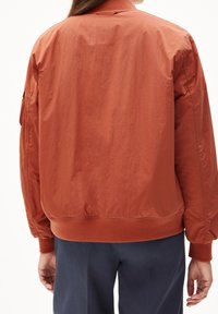 Giacca bomber color ruggine con tessuto leggero e testurizzato, polsini e cintura a costine, e design minimalista. Non presenta elementi metallici visibili.