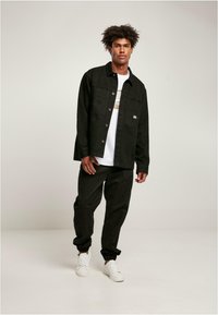 Svart denimskjortejakke med knapper, brystlomme og hvit logo på en hvit t-skjorte. Svart joggebukse og hvite sneakers.