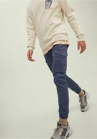 Osoba oblečená v béžovém svetru, pruhované košili, modrých cargo kalhotách a béžových teniskách, kráčející na jednolitém pozadí.