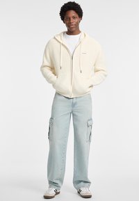 Kremeni rebrasti zip-up hoodie s kapuljačom na vezanje, uparen s svijetlopPlavim cargo hlačama koje imaju bočne džepove i bijelim tenisicama.