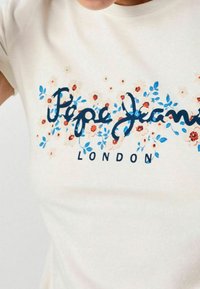Světle krémové tričko s květinovými akcenty a nápisem "Pepe Jeans LONDON" v námořnické modré. Krátké rukávy a kulatý výstřih.