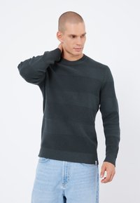 Pull en maille vert foncé avec des rayures texturées horizontales, un col rond et des poignets côtelés. Associé à un short en denim bleu clair.
