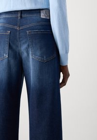 Oseba v temno modri denim hlačah z zadnjimi žepi in svetlo modrem dolgih rokavih topu, ki stoji ob enotni ozadju.