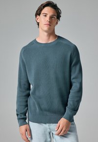 Pull en tricot côtelé bleu foncé avec un col rond et des manches longues, présentant un ourlet droit et un tissu texturé. Assorti à un jean clair.