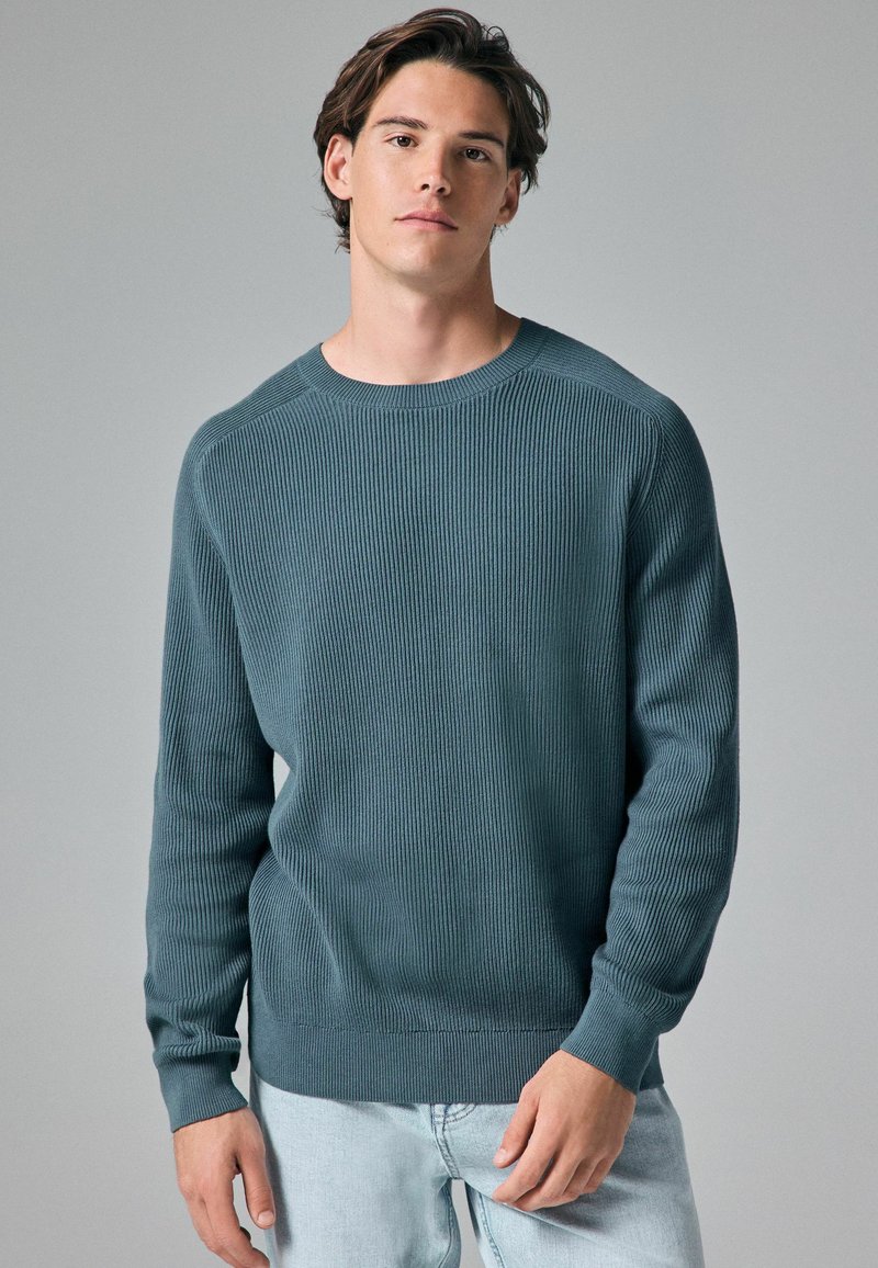 Pull en tricot côtelé bleu foncé avec un col rond et des manches longues, présentant un ourlet droit et un tissu texturé. Assorti à un jean clair.