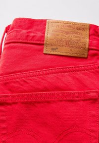 Tissu en denim rouge avec une patch en cuir. La patch présente un texte et un design en relief, indiquant les détails de la marque et du produit. Comprend des accents de couture.