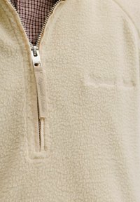 Hellbeiger Fleecepullover mit Viertel-Reißverschluss, einer strukturierten Oberfläche und einem dezenten auf der Brust gestickten Logo. Karomuster-Futter sichtbar.