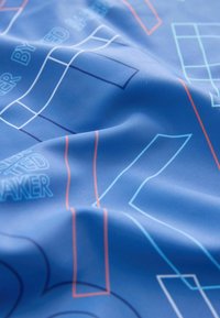 Blåt stof med geometriske former i hvid, lyseblå og orange linjer samt delvist tekst, vist med bløde folder og teksturdetaljer.