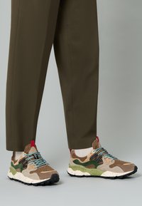 Las zapatillas multicolores presentan una mezcla de materiales en marrón, verde y beige con un patrón texturizado, cordones azules y una suela salpicada en negro y blanco.