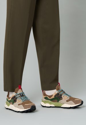 Des baskets multicolores présentent un mélange de matériaux marron, vert et beige avec un motif texturé, des lacets bleus et une semelle mouchetée noire et blanche.