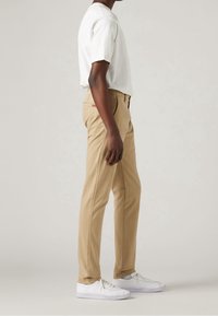 Persona che indossa pantaloni beige a gamba dritta, una camicia bianca a maniche corte e sneakers bianche, in piedi di lato contro uno sfondo semplice.
