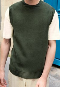 Homme portant un gilet sans manches en tricot vert foncé sur une chemise à manches courtes crème, debout à l'extérieur près d'une porte bleue.