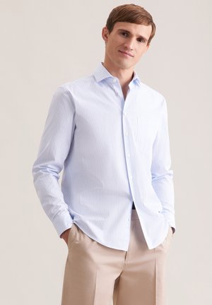 Giovane uomo con capelli castano chiaro che indossa una camicia azzurra a righe con bottoni e pantaloni beige, mani in tasca, sorriso leggero.