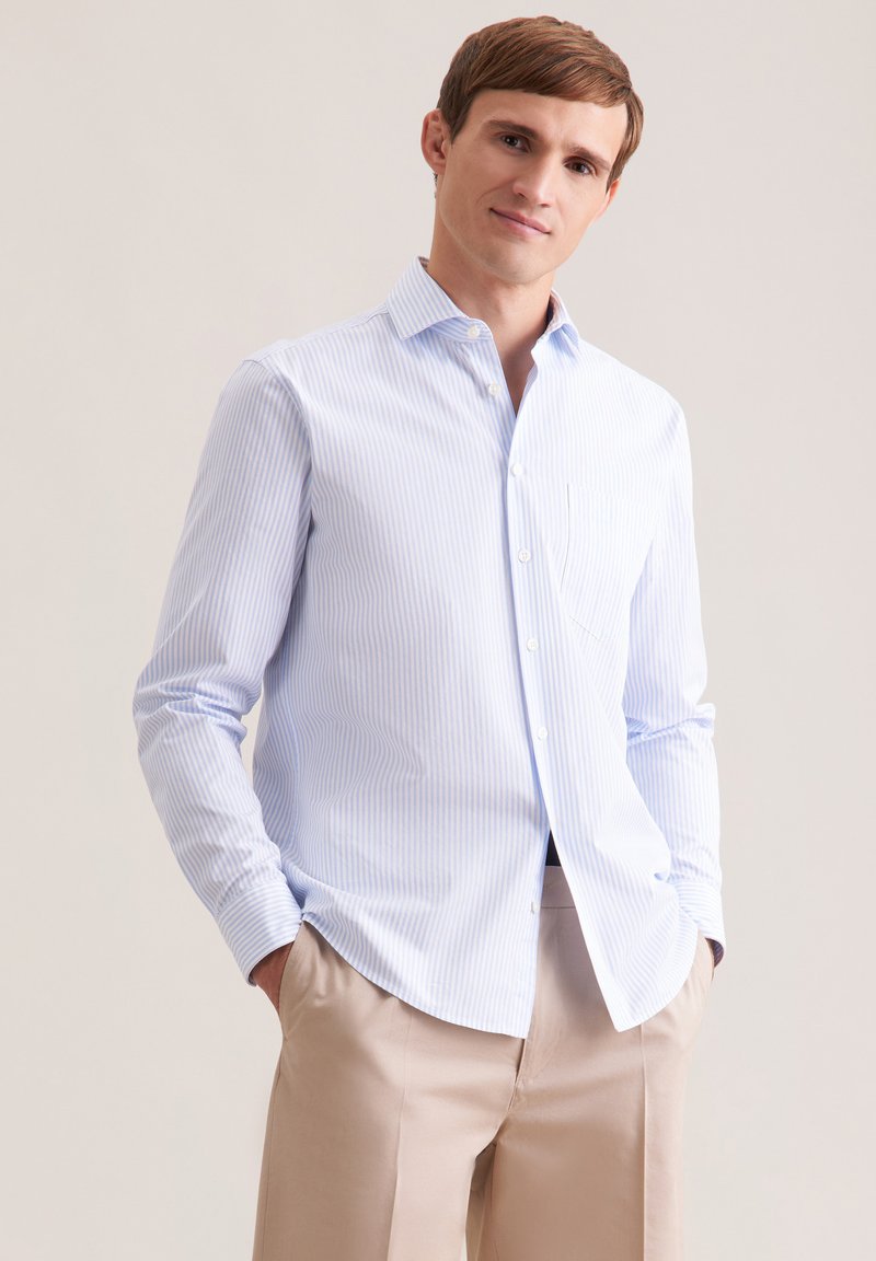 Jeune homme aux cheveux châtain clair portant une chemise à rayures bleues claires boutonnée et un pantalon beige, mains dans les poches, souriant légèrement.