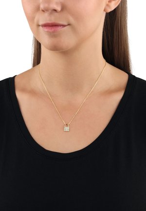 Femme portant un collier en chaîne dorée avec un petit pendentif en forme de cadenas doré, habillée d'un haut noir à décolleté en scoop, sur fond blanc.
