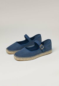 Alpargatas planas de color azul marino con punta cerrada, detalle de correa y hebilla, y suela de cuerda de yute natural. Textura suave de ante.