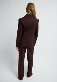 Persona con cabello largo que lleva un traje de raya diplomática marrón oscuro y zapatos beige de punta, de pie frente a un fondo claro y liso.
