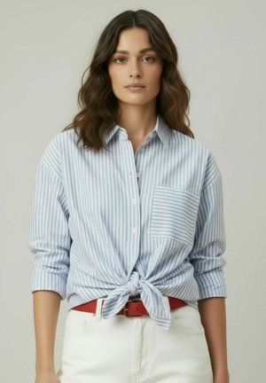 Femme aux cheveux bruns ondulés portant une chemise à rayures bleues et blanches nouée à la taille, un pantalon blanc et une ceinture rouge, faisant face à l'avant.