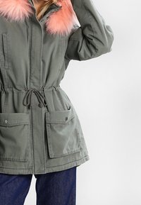 Olivgrüner Parka mit einem rosa Fellkragen, Zugband in der Taille und zwei Fronttaschen. Hergestellt aus einem strapazierfähigen, weichen Stoff.