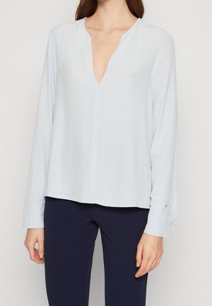 Femme portant un chemisier bleu clair à manches longues avec un col en V et un pantalon bleu foncé, debout devant un fond uni.