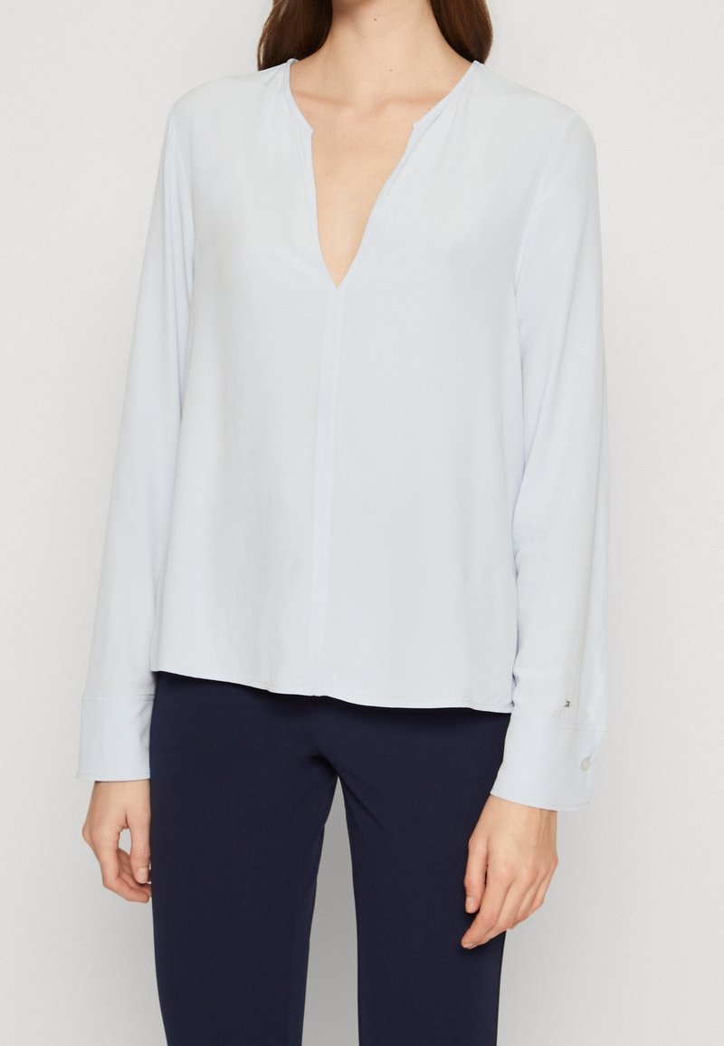 Femme portant un chemisier bleu clair à manches longues avec un col en V et un pantalon bleu foncé, debout devant un fond uni.