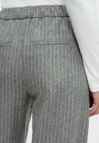 Prim-plan al unei persoane care poartă pantaloni gri cu dungi subțiri și buzunare cu deschidere pe partea din spate, purtând un top alb cu mâneci lungi, cu mâna sprijinită pe lângă corp.