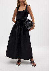 Top negro sin mangas con escote cuadrado, combinado con una falda negra larga y fluida. Sosteniendo un bolso negro estructurado y usando sandalias negras planas.