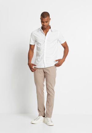 Camicia bianca a maniche corte con colletto a punta, abbinata a pantaloni a righe beige chiaro e sneaker bianche. Design minimale, senza fantasie.