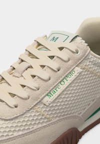 Κοντινή λήψη μπεζ αθλητικού παπουτσιού με υφή κυψελίδας, κρέμ κορδόνια και πράσινο λογότυπο "Marc O'Polo" στο πλάι και στη γλώσσα.