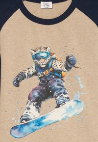 Snehová leopardka v okuliaroch a zimnom oblečení, snowboarduje s modrým snowboardom a rozprášeným snehom na béžovej košeli.