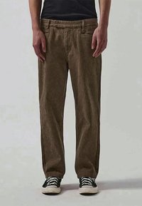 Calças castanhas, texturizadas, com um corte relaxado. Apresenta dois bolsos frontais e costuras laterais. Combinadas com ténis de atacadores em preto e branco.