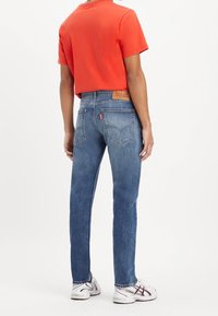 Jeans de mezclilla azul ajustados con un parche de cuero en la cinturilla trasera, combinados con una camiseta roja de manga corta y zapatillas blancas.