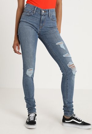 Personne portant un jean skinny bleu déchiré, des baskets noires et blanches, et un haut orange, debout avec une main détendue le long du corps.