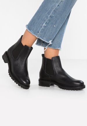 Schwarze Chelsea-Lederstiefel mit elastischen Seitenteilen, getragen zu gekürzten, ausgefransten blauen Jeans, gezeigt bei gekreuzten Beinen vor weißem Hintergrund.