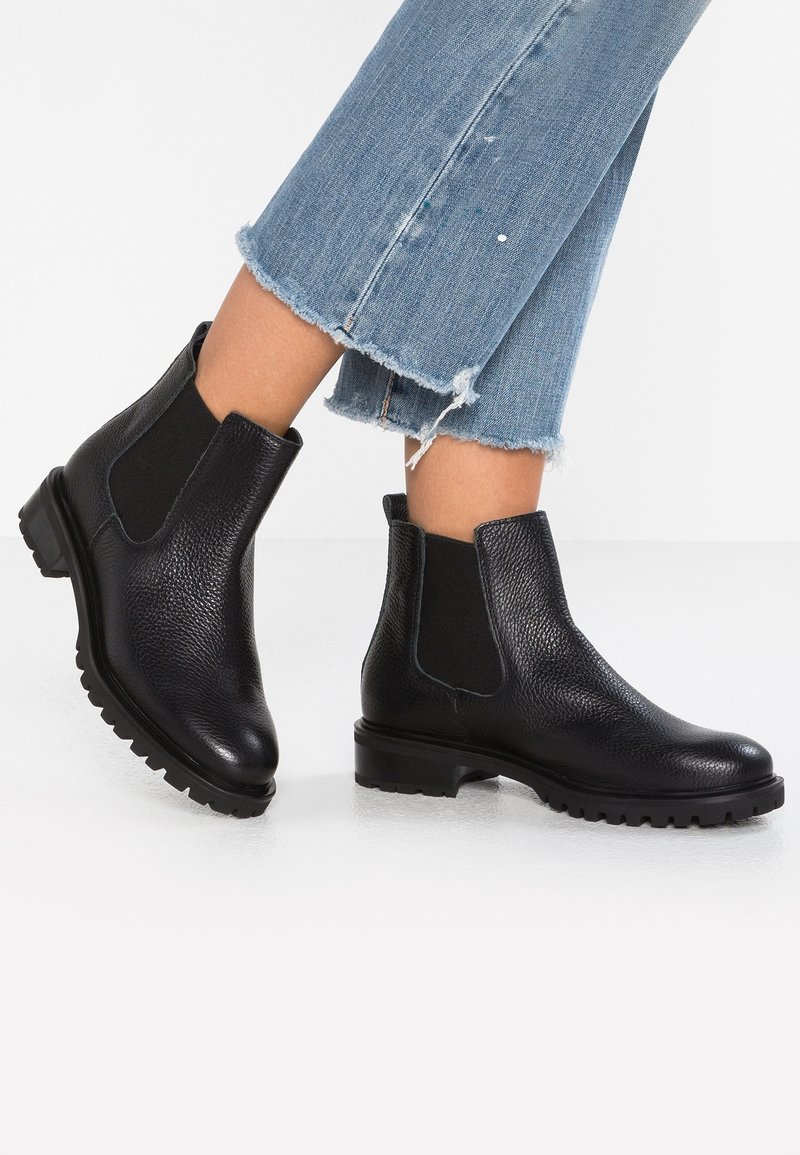 Bottes Chelsea en cuir noir avec panneaux latéraux élastiques portées avec un jean bleu raccourci et effiloché, montrées sur des jambes croisées sur fond blanc.