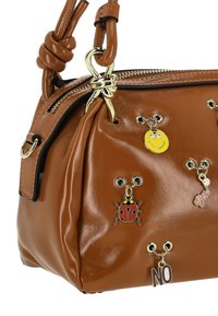 Barna műbőr kézitáska lekerekített formával, csomózott pánttal és fém medálokkal, köztük egy katica és egy mosolygós arc.