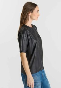 Donna con lunghi capelli castani, indossa una maglietta nera lucida a maniche corte e jeans blu, in piedi di profilo su uno sfondo semplice.